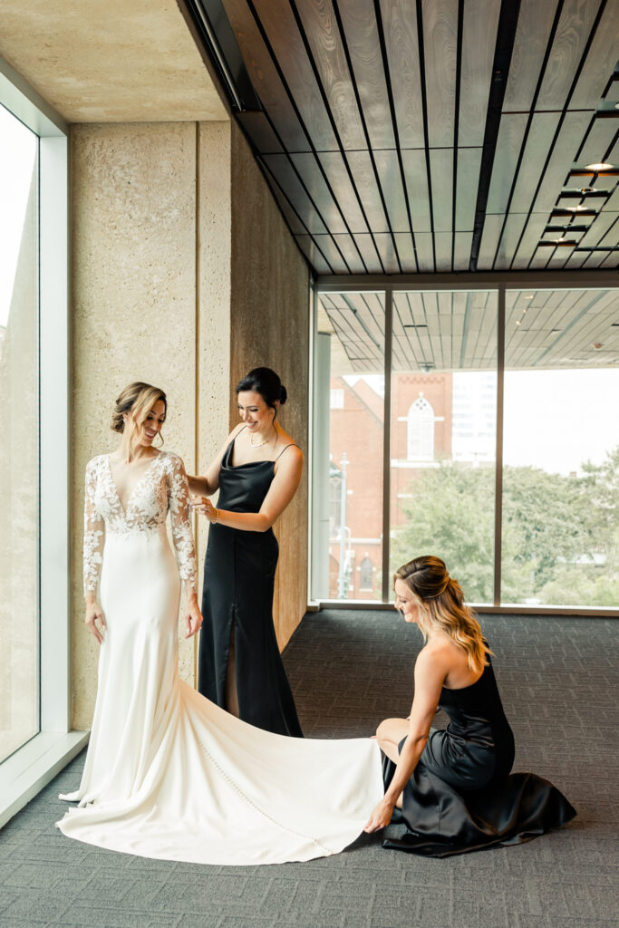 Bride, in white long sleeve lace dress, getting ready with two sisters at the Mint Museum wedding venue in Uptown Charlotte
