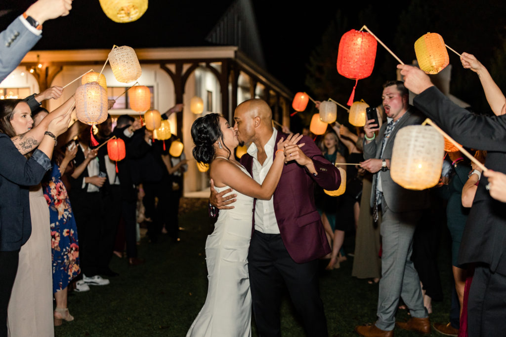 Bride, in white dress, kissing groom, in burgundy suit, under Chinese lanterns during wedding exit Bride and groom first kiss during ceremony at the Bradford wedding venue in Raleigh NC.