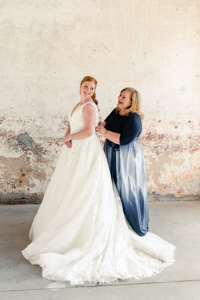 Mother of bride, in a blue dress, zips up the Bride's white wedding dress at Providence Cotton Mill wedding venue. Photographed by Charlotte wedding photographer, Stephanie Bailey Photography.