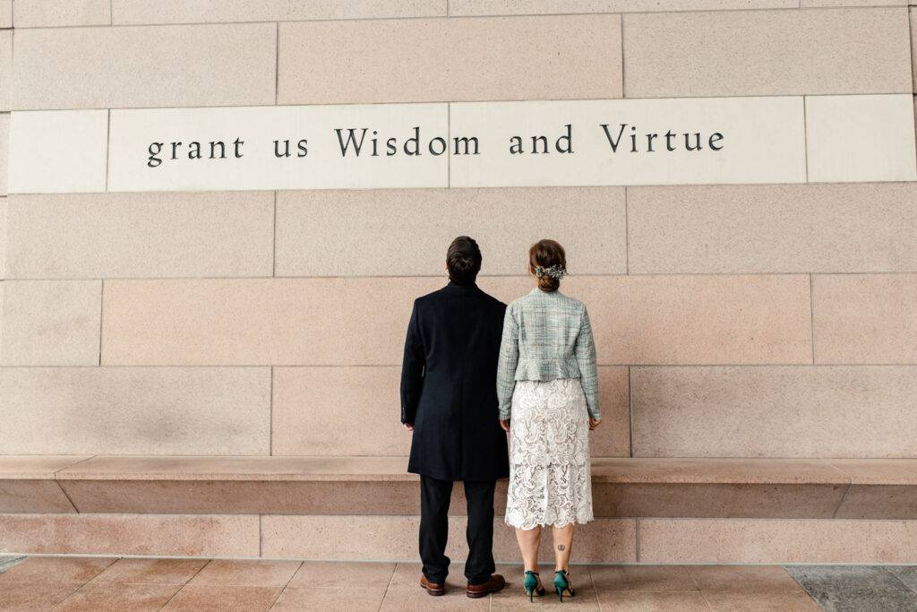 Bride and groom looking up at writing on the wall that states, "grant us wisdom and virtue during their courthouse elopement in Uptown Charlotte. Photographed by Stephanie Bailey Photography. 