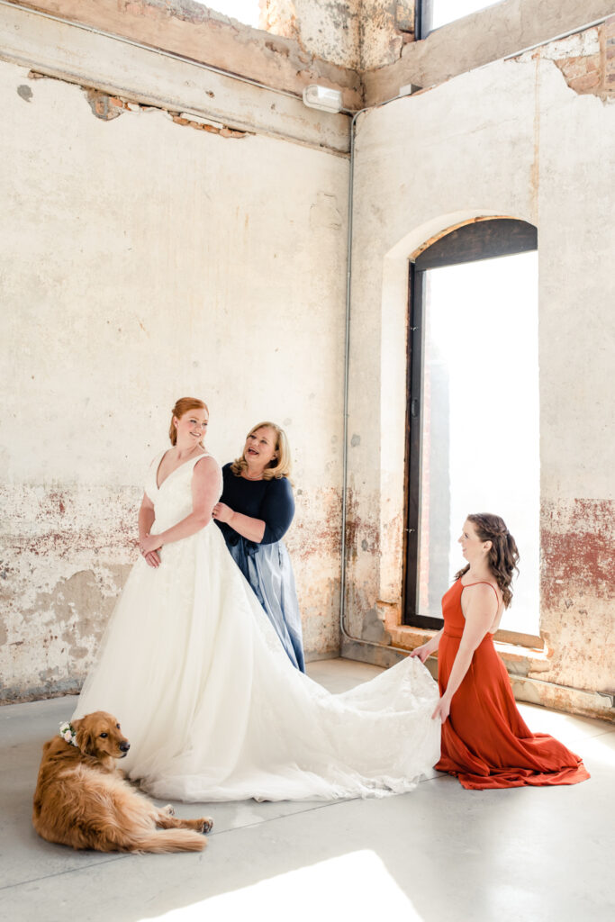 Mother of bride, in a blue dress, zips up the Bride's white wedding dress. Bride's sister, in a orange dress, adjusts the train of the Bride's dress. Golden retriever dog sits by the bride at Providence Cotton Mill wedding venue. Photographed by Charlotte wedding photographer, Stephanie Bailey Photography.