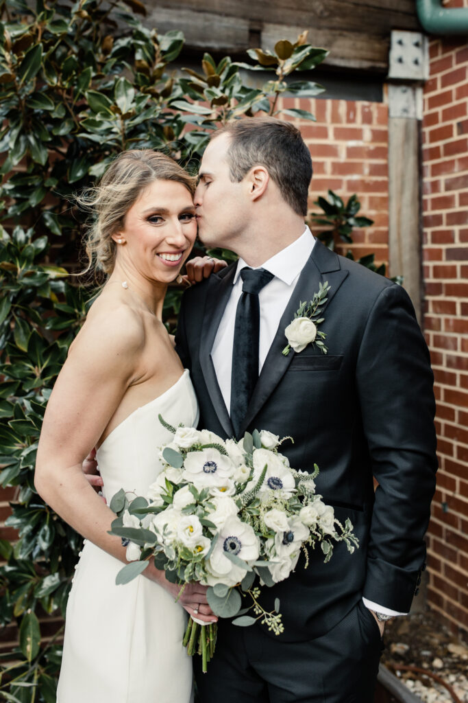 Bride in white dress smiling while groom in black suit kisses her cheek at Byron's south end wedding venue. Photographed by Charlotte wedding photographer. 
