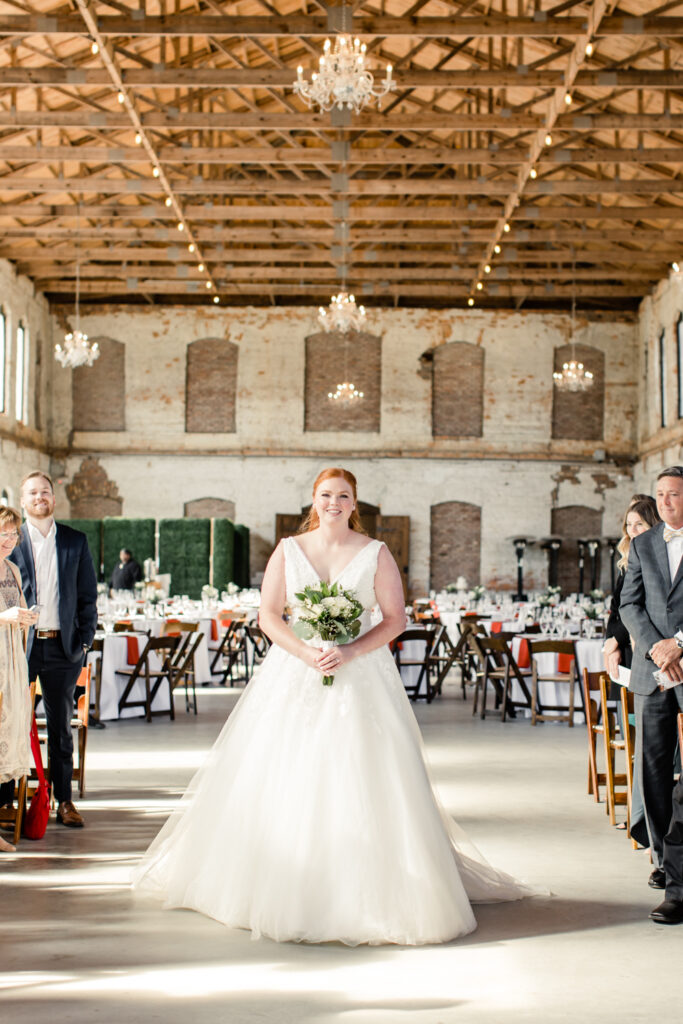 Bride, in white dress holding white and green bouquet, walks down ceremony aisle at Providence Cotton Mill wedding venue. Photographed by Charlotte wedding photographer, Stephanie Bailey Photography.