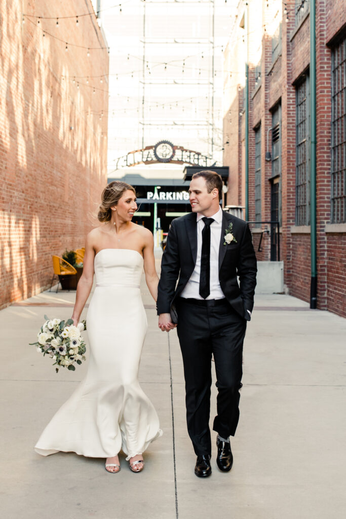 Bride in white dress smiling holding hands with groom in black suit walking at Byron's south end wedding venue. Photographed by Charlotte wedding photographer. 