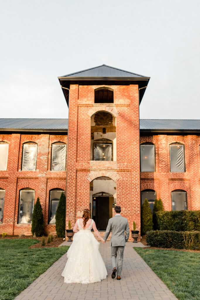 Bride, in white gown, holding hands with groom, in grey suit, walk toward the Providence Cotton Mill wedding venue. Photographed by Charlotte wedding photographer, Stephanie Bailey Photography.