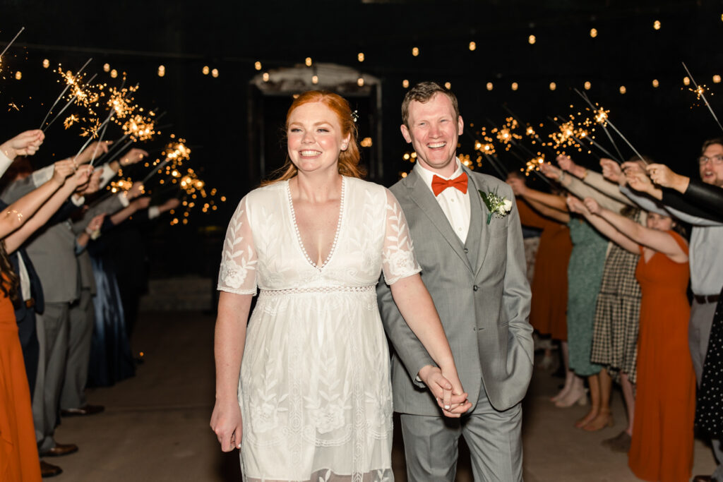 Bride, in white dress and cowboy boots, holding hands with groom, in grey suit and orange bow tie, walk under sparklers during wedding exit at Providence Cotton Mill wedding venue. Photographed by Charlotte wedding photographer, Stephanie Bailey Photography.