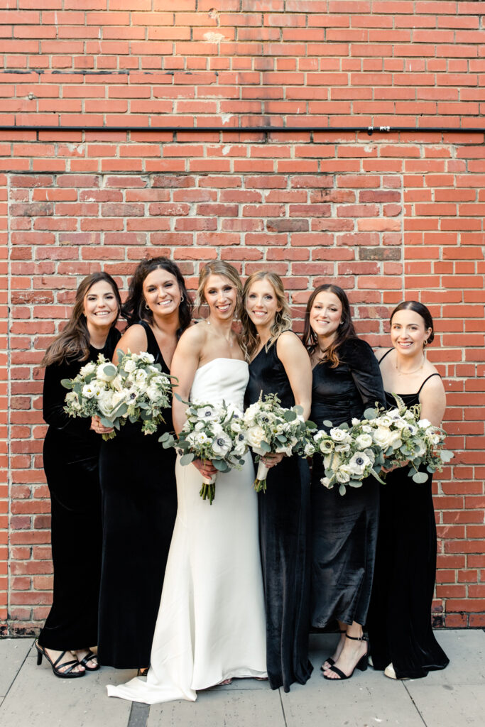 Bride, in white dress, and bridesmaids, in black dresses, hold white bouquets in front of red brick wall at Byron's South End wedding venue. Photographed by Charlotte wedding photographer, Stephanie Bailey Photography.