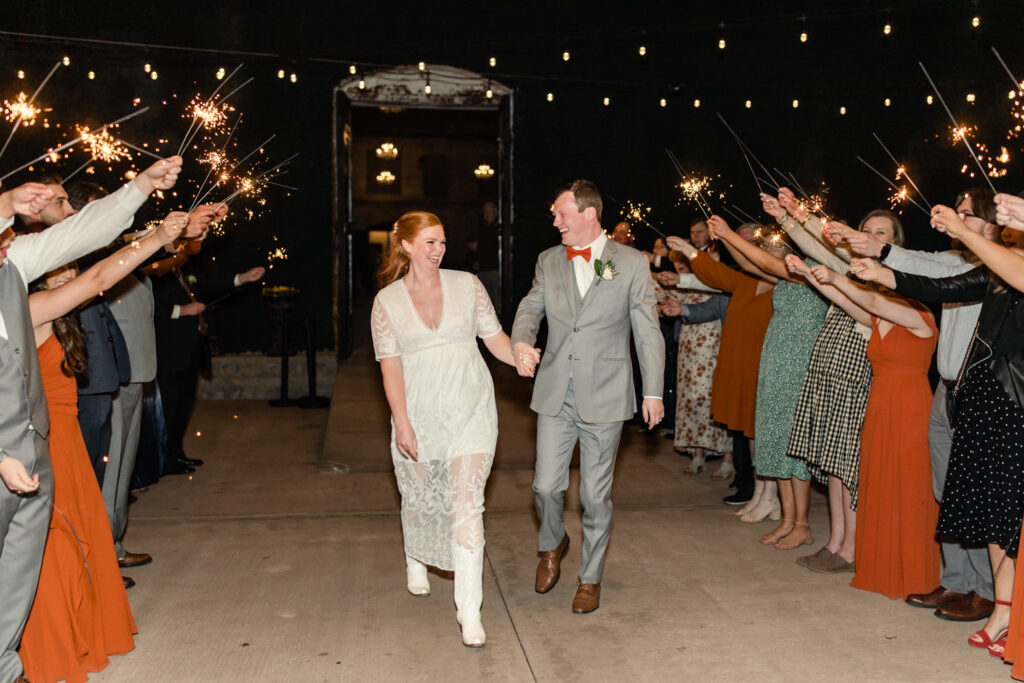 Bride, in white dress and cowboy boots, holding hands with groom, in grey suit and orange bow tie, walk under sparklers during wedding exit at Providence Cotton Mill wedding venue. Photographed by Charlotte wedding photographer, Stephanie Bailey Photography.