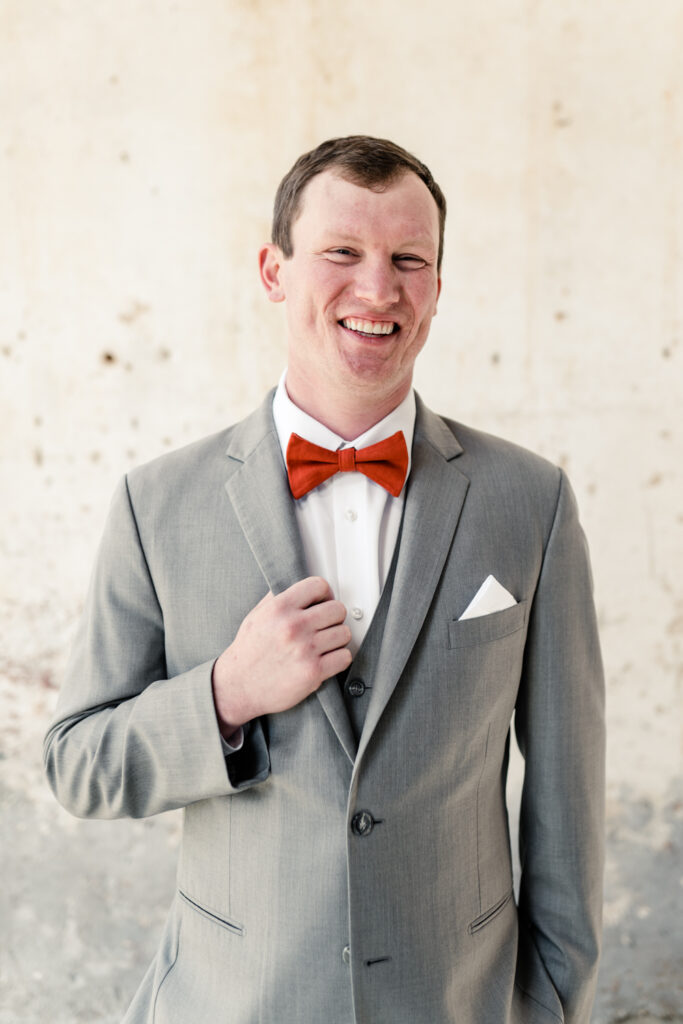 Groom wearing a grey suit and orange bow tie smiles at Providence Cotton Mill wedding venue. Photographed by Charlotte wedding photographer, Stephanie Bailey Photography.