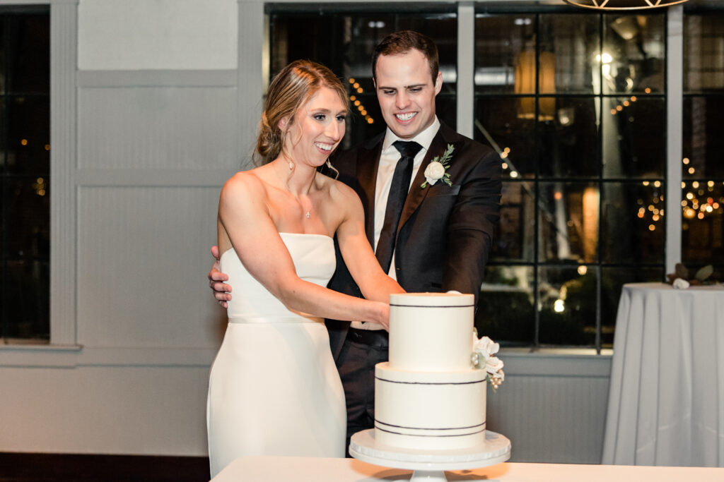 Bride, in strapless white dress, cuts two tiered white cake with groom, in black suit, at Byron's South End wedding venue. Photographed by Charlotte wedding photographer, Stephanie Bailey Photography.