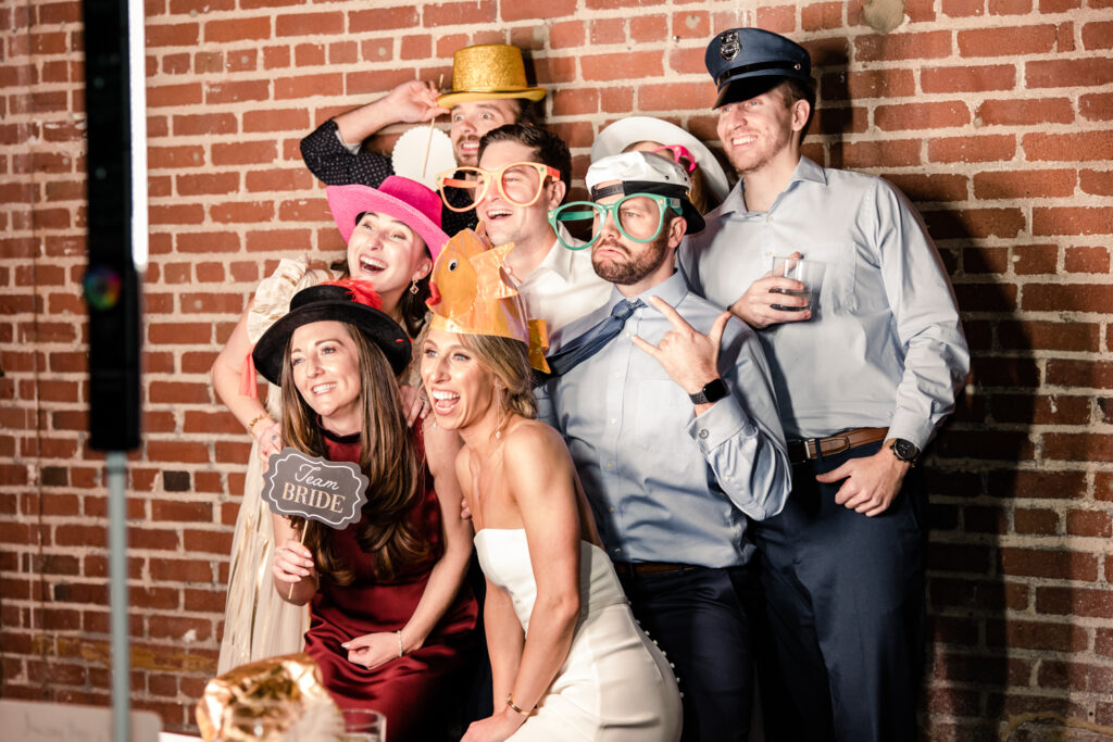 Bride, in white dress, and 6 wedding guests pose in front of photo booth wearing silly hats at Byron's South End wedding venue. Photographed by Charlotte wedding photographer, Stephanie Bailey Photography.