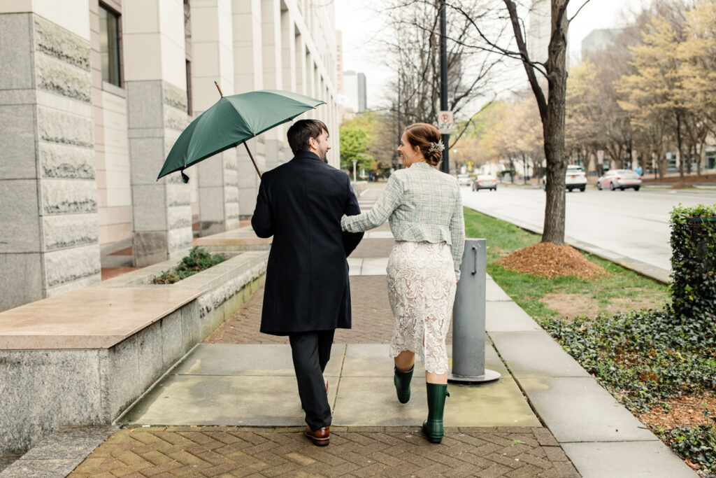 Bride and groom walking in the rain, holding a green umbrella after their courthouse elopement in Uptown Charlotte. Photographed by Stephanie Bailey Photography. 