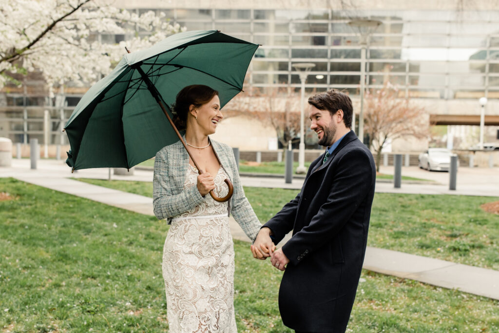 Bride and groom walking in the rain, holding a green umbrella after their courthouse elopement in Uptown Charlotte. Photographed by Stephanie Bailey Photography. 