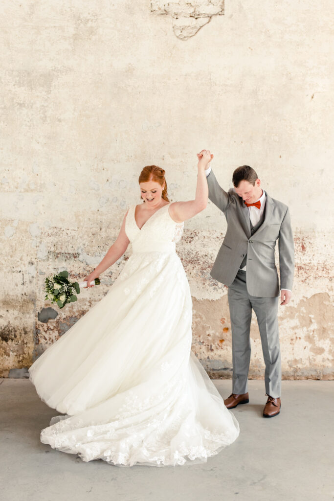 Groom, in grey suit and orange bow tie, twirls bride, in white gown at Providence Cotton Mill wedding venue. Photographed by Charlotte wedding photographer, Stephanie Bailey Photography.