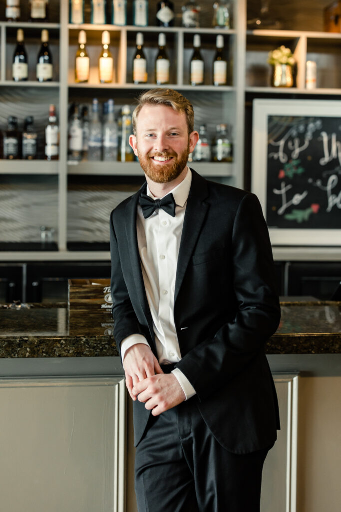 Groom, in black suit and bow tie, leaning on a bar smiling at 1705 East Raleigh Wedding Venue. Photographed by Charlotte Wedding Photographer, Stephanie Bailey Photography.
