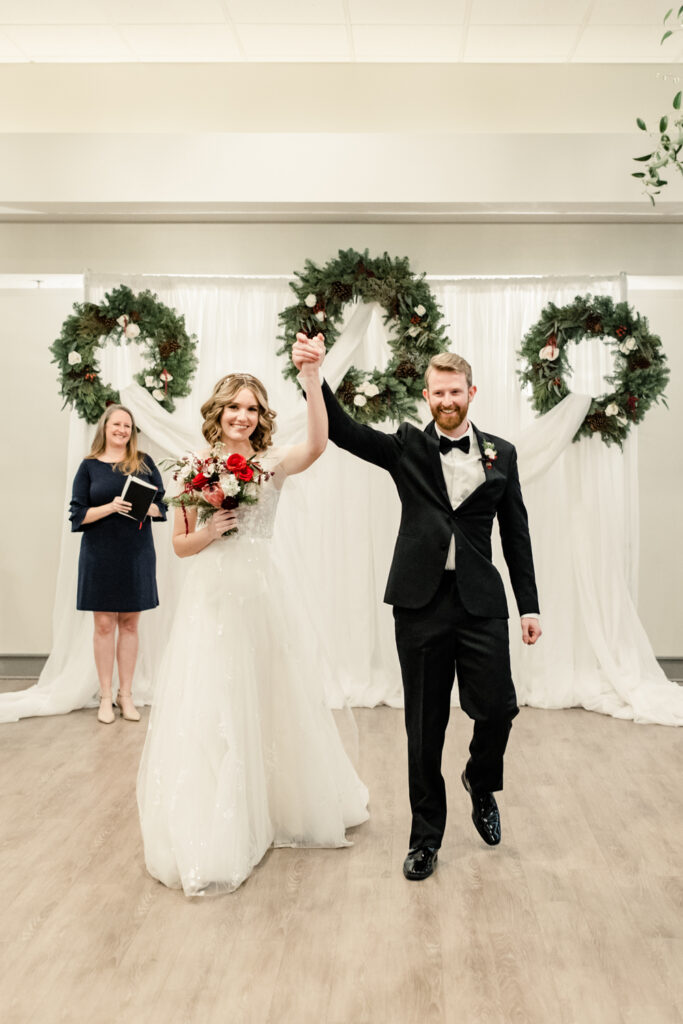 Bride, in white dress, holding hands with groom, in black suit with red floral boutonnière during ceremony at 1705 East Raleigh Wedding Venue. Photographed by Charlotte Wedding Photographer, Stephanie Bailey Photography.