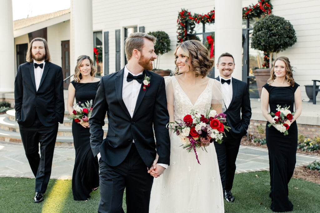 Groom, in black suit and bow tie, walking and holding hands with bride, in white gown followed by wedding party at 1705 East Raleigh Wedding Venue. Photographed by Charlotte Wedding Photographer, Stephanie Bailey Photography.