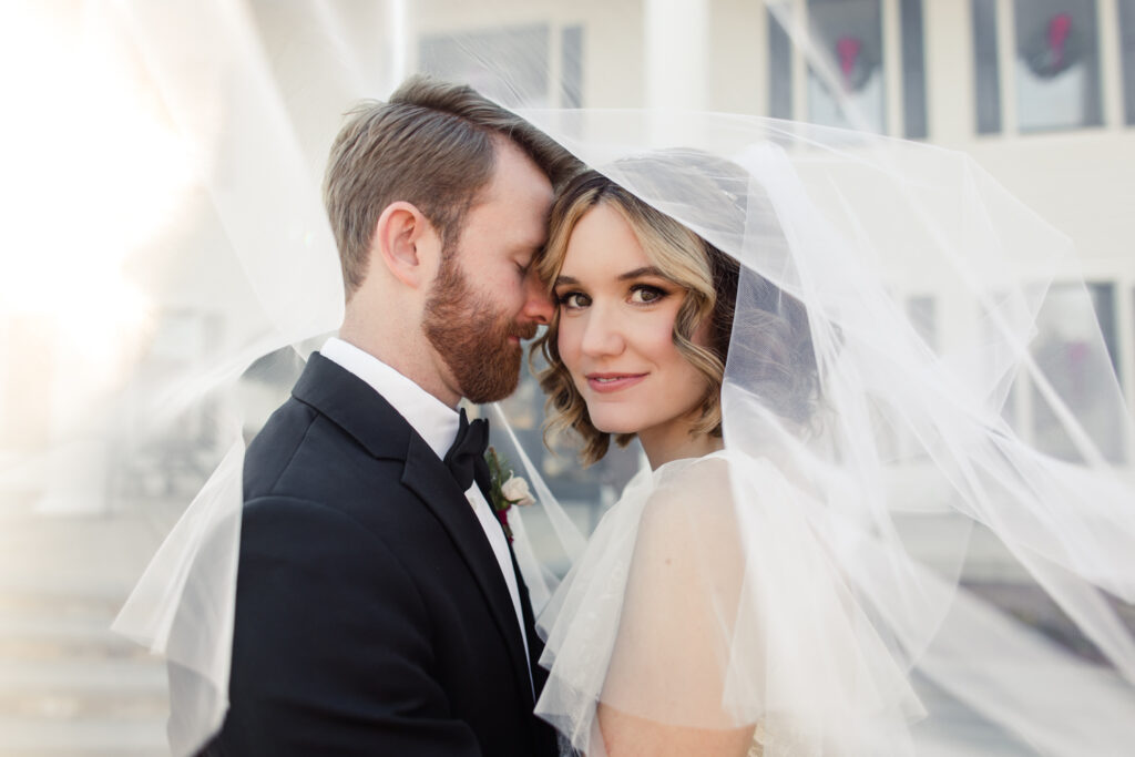 Bride, in white gown and veil, smiling as groom, in black suit, nuzzles on her cheek at 1705 East Raleigh Wedding Venue. Photographed by Charlotte Wedding Photographer, Stephanie Bailey Photography.