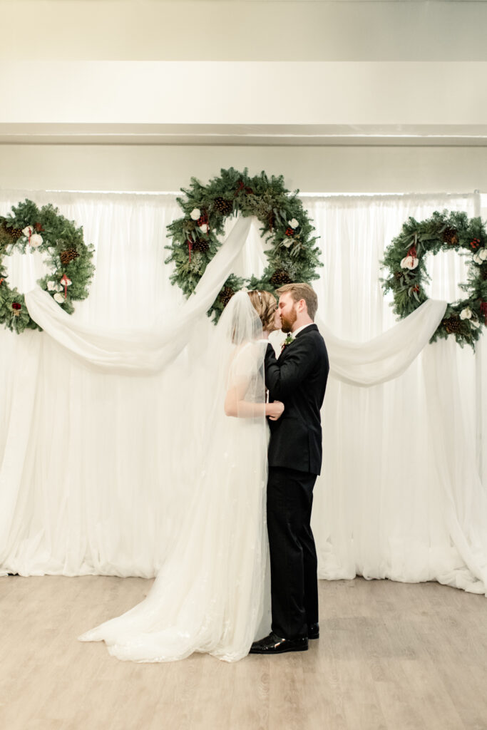 Bride, in white gown and veil, kissing her groom, in black suit under Christmas wreaths at 1705 East Raleigh Wedding Venue. Photographed by Charlotte Wedding Photographer, Stephanie Bailey Photography.