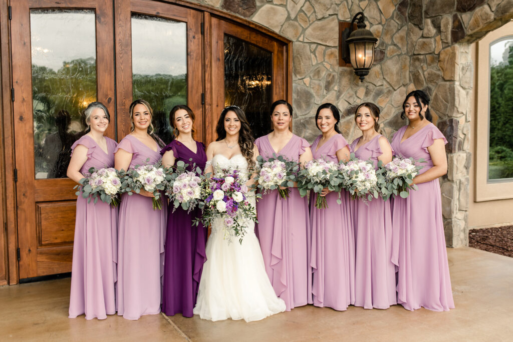 Bride, with long brown hair wearing white, long laced wedding dress, holds a purple and white bouquet of flowers standing with seven bridesmaids, in purple dresses and flowers, in front of brown stone wall Tuscan Ridge Wedding Venue near Charlotte NC