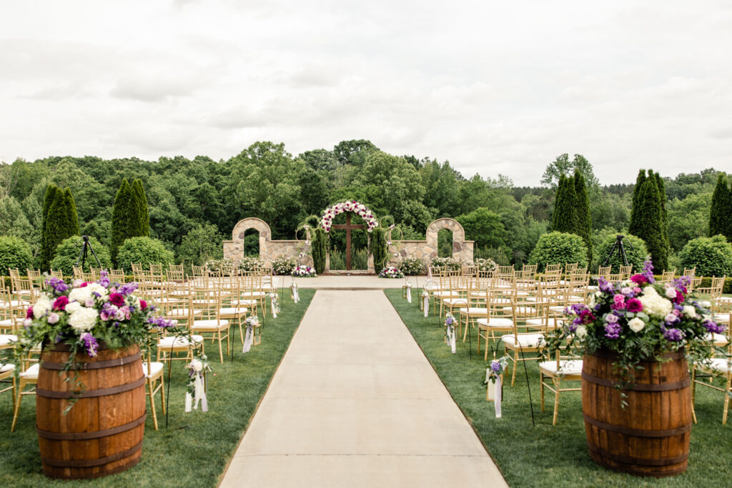 Outdoor wedding ceremony with grass lawn and pavement aisle. Purple and white flowers on brown barrels on both sides. There are three stone arches decorated with purple floral arrangements at Tuscan Ridge Wedding Venue near Charlotte NC