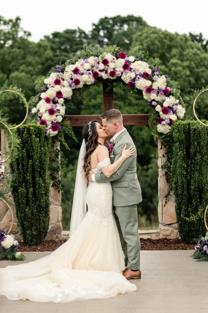 Bride, in white lace dress with long brown hair, kisses groom, in green suit with short brown hair, in front of wooden cross with purple flowers at Tuscan Ridge Wedding Venue near Charlotte NC