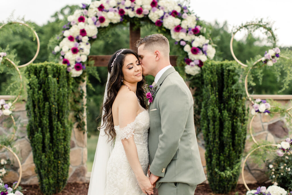 Groom, in green suit with short brown hair, kisses the cheek of bride, in white lace dress with long brown hair, in front of wooden cross with purple flowers at Tuscan Ridge Wedding Venue near Charlotte NC