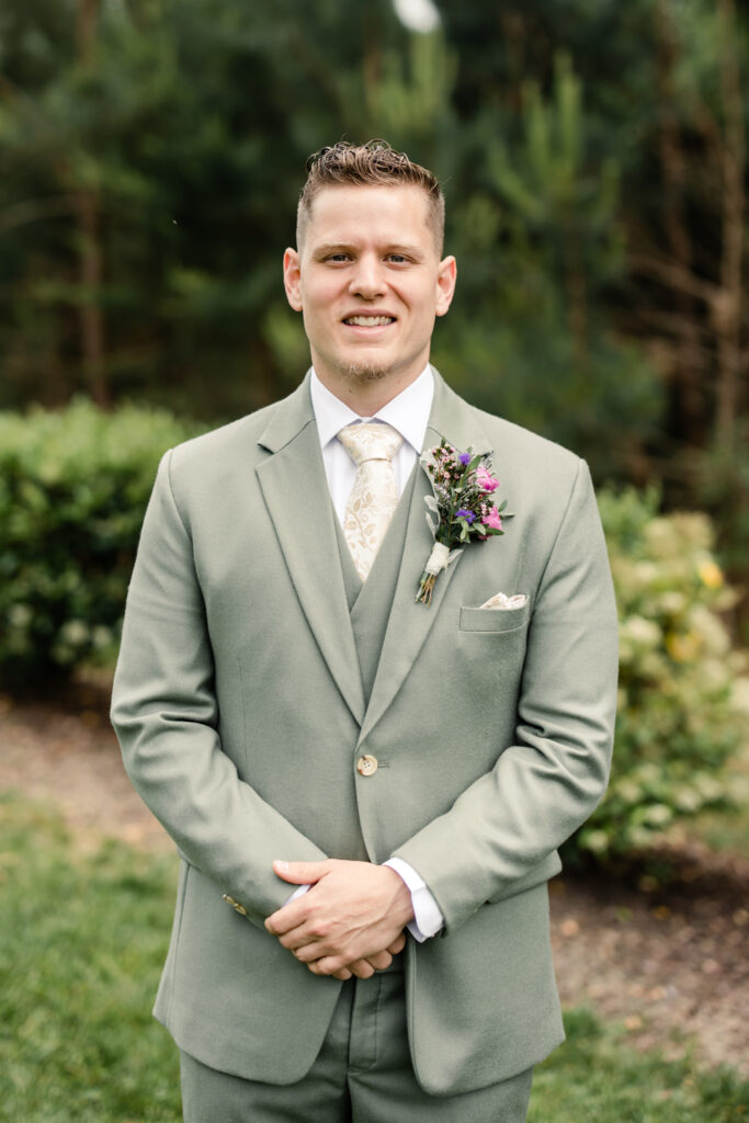 Groom, with brown hair wearing green suit and beige tie with pink and purple boutonnière smiling in front of green trees at Tuscan Ridge Wedding Venue near Charlotte NC. 