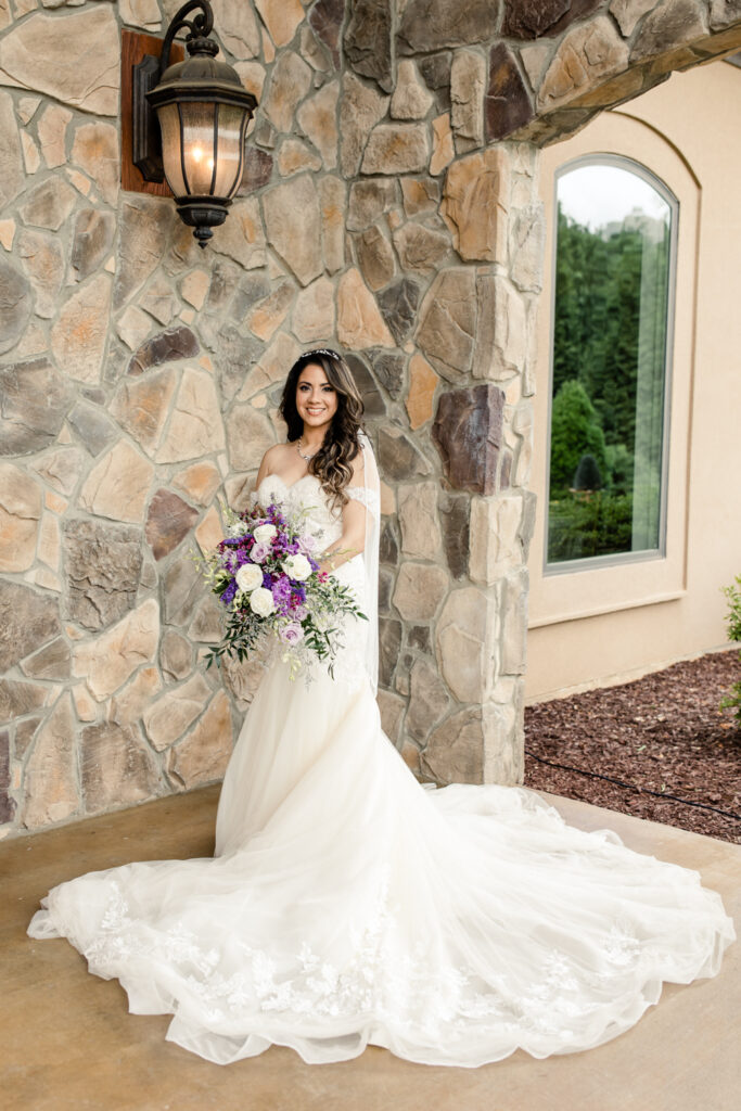 Bride, with long brown hair wearing white, long laced wedding dress, holds a purple and white bouquet of flowers in front of brown stone wall Tuscan Ridge Wedding Venue near Charlotte NC