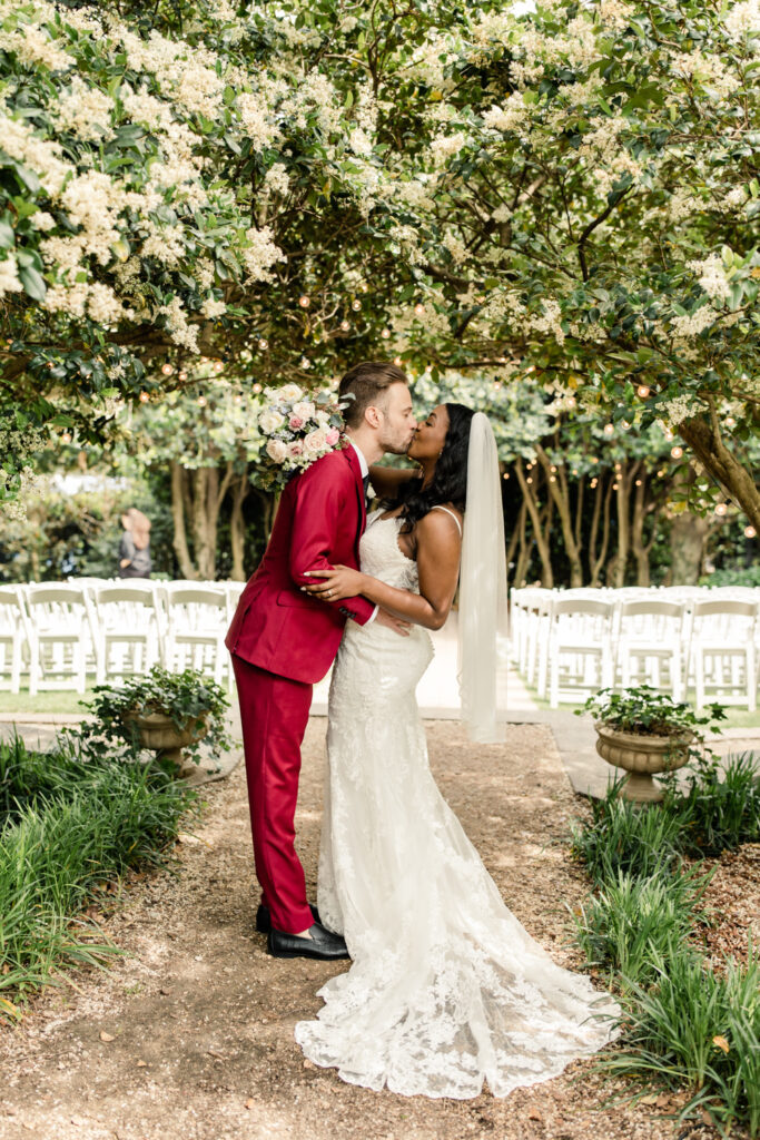 Bride, in white gown, kissing groom, in red suit, under a canopy of greenery at McGill Rose Garden in Charlotte NC.