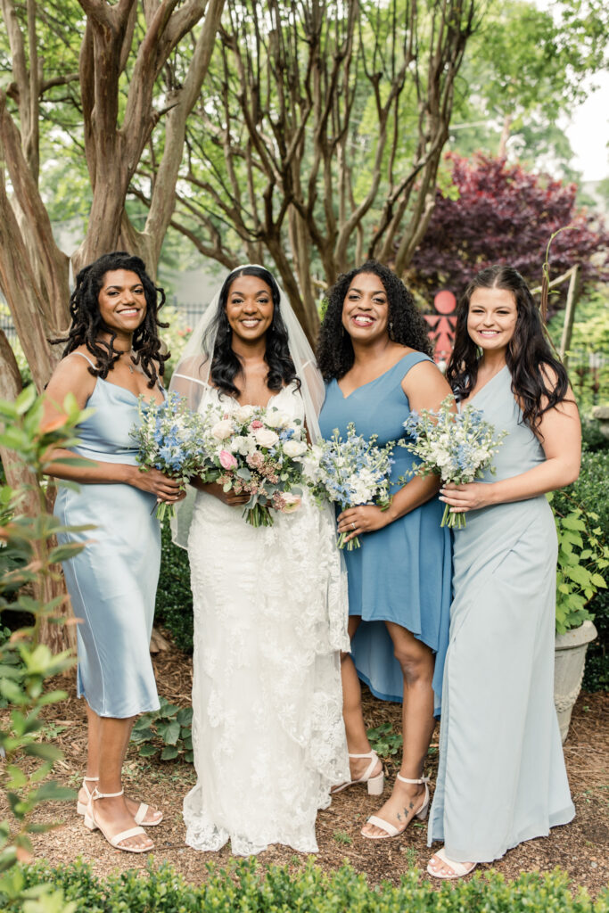 Bride, in white gown holding green and white flowers, standing with three bridesmaids in blue dresses and flowers at McGill Rose Garden in Charlotte NC.