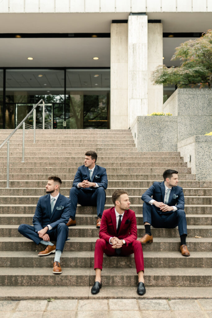 Groom, in red suit, sitting on outdoor staircase in uptown Charlotte with three groomsmen in blue suits.