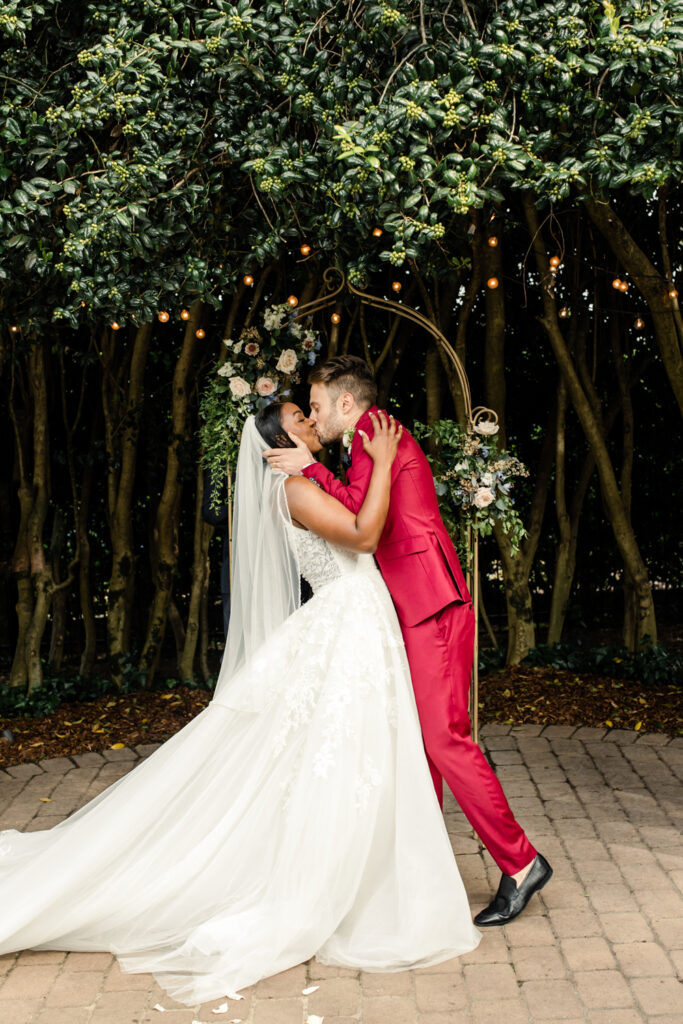Bride, in white gown, kissing groom, in red suit, under a canopy of greenery during ceremony at McGill Rose Garden in Charlotte NC.