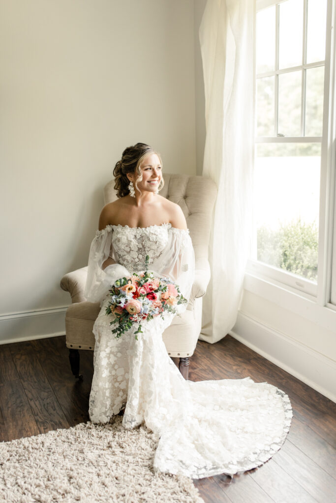 Bride, with brown hair in pony tail, sits on cream chair looking out window holding pink floral bouquet at Morning Glory Farm Wedding Venue in Charlotte NC.