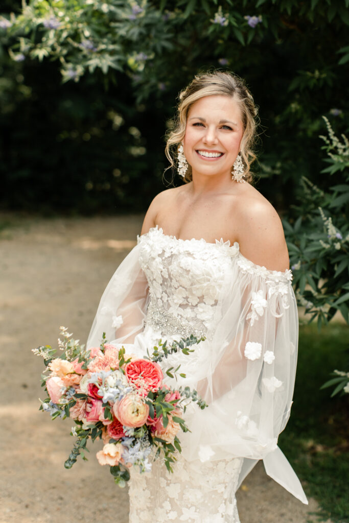 Bride, with brown hair in pony tail, smiles holding pink floral bouquet at Morning Glory Farm Wedding Venue in Charlotte NC.