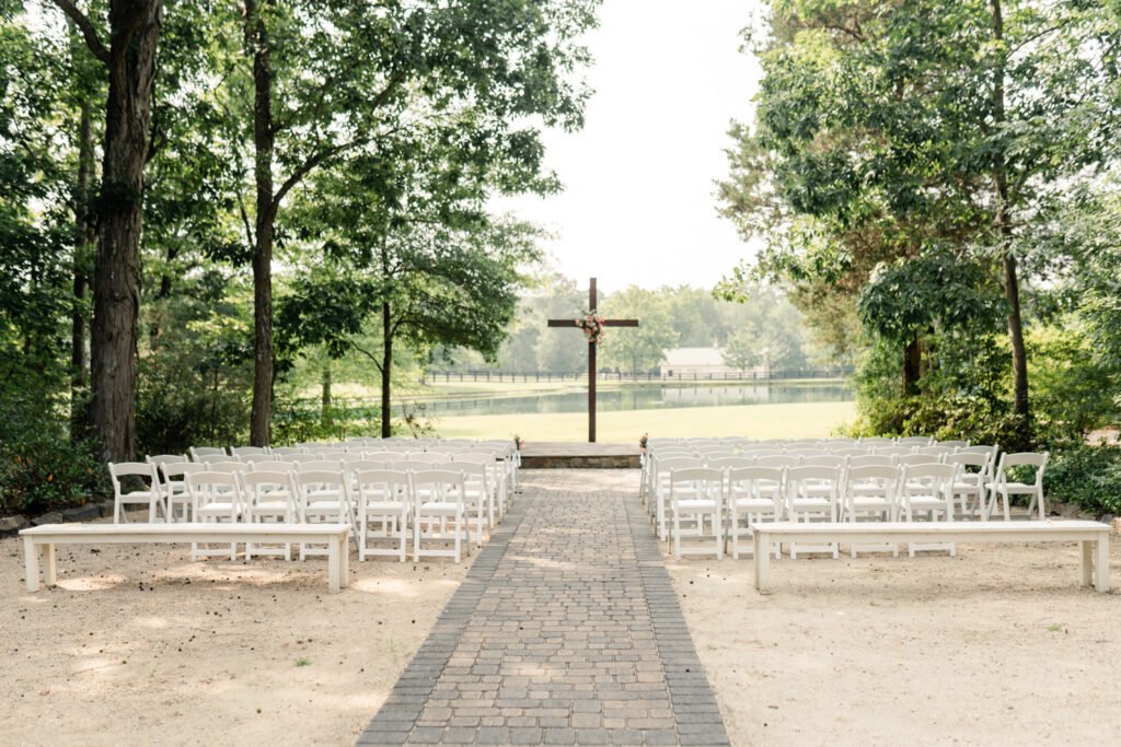 Outdoor wedding ceremony in front of pond in the woods with white chairs and wooden cross at Morning Glory Farm Wedding Venue in Charlotte NC