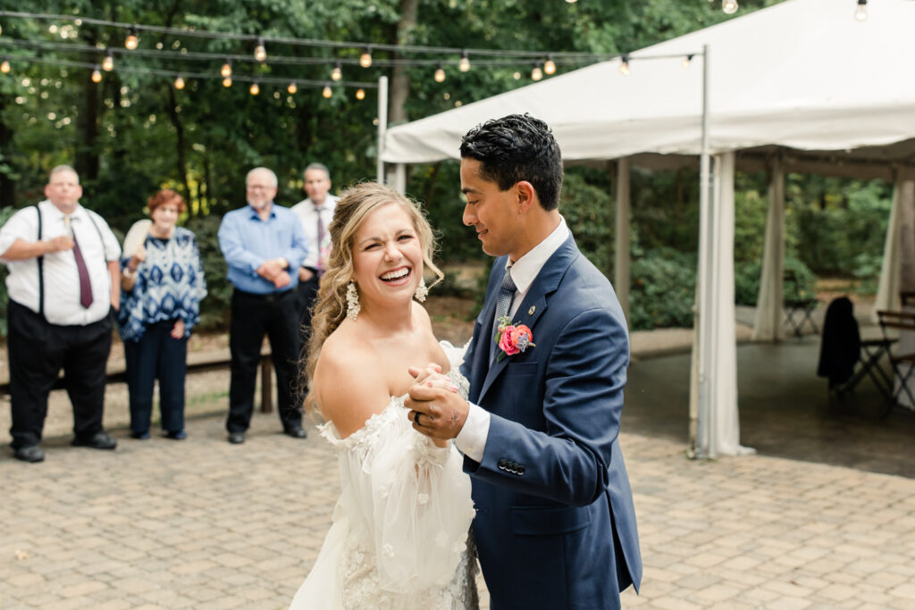 Bride, in white lace dress, slow dances with groom, in blue suit with pink boutonniere during first dance at Morning Glory Farm Wedding Venue in Charlotte NC.