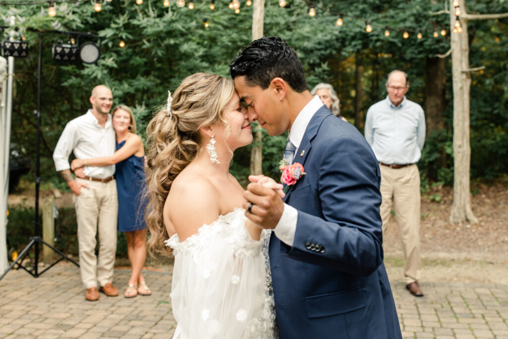 Bride, in white lace dress, nose to nose with groom, in blue suit with pink boutonniere during first dance at Morning Glory Farm Wedding Venue in Charlotte NC.