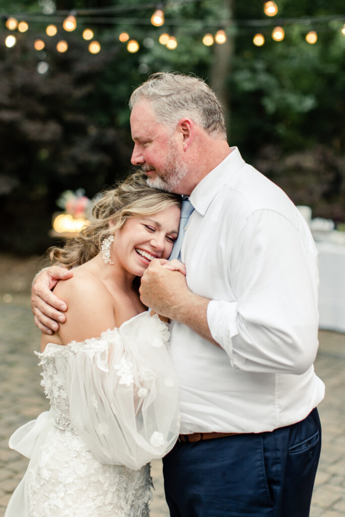 Bride, in white lace dress with long sleeves, lays her head on father's chest during first dance at Morning Glory Farm Wedding Venue in Charlotte NC.