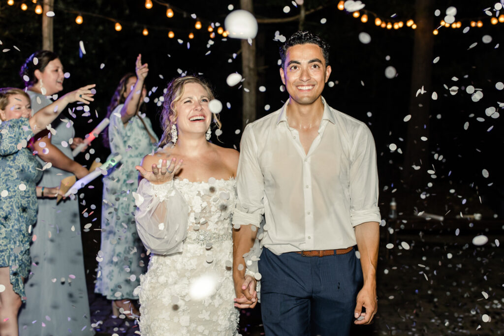 Bride, in white lace dress, holds hands with groom, in white button down and blue pants, walking under confetti at night during reception exit at Morning Glory Farm Wedding Venue in Charlotte NC.