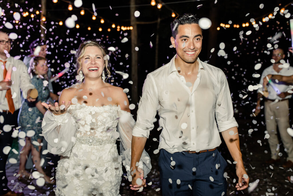 Bride, in white lace dress, holds hands with groom, in white button down and blue pants, walking under confetti at night during reception exit at Morning Glory Farm Wedding Venue in Charlotte NC.