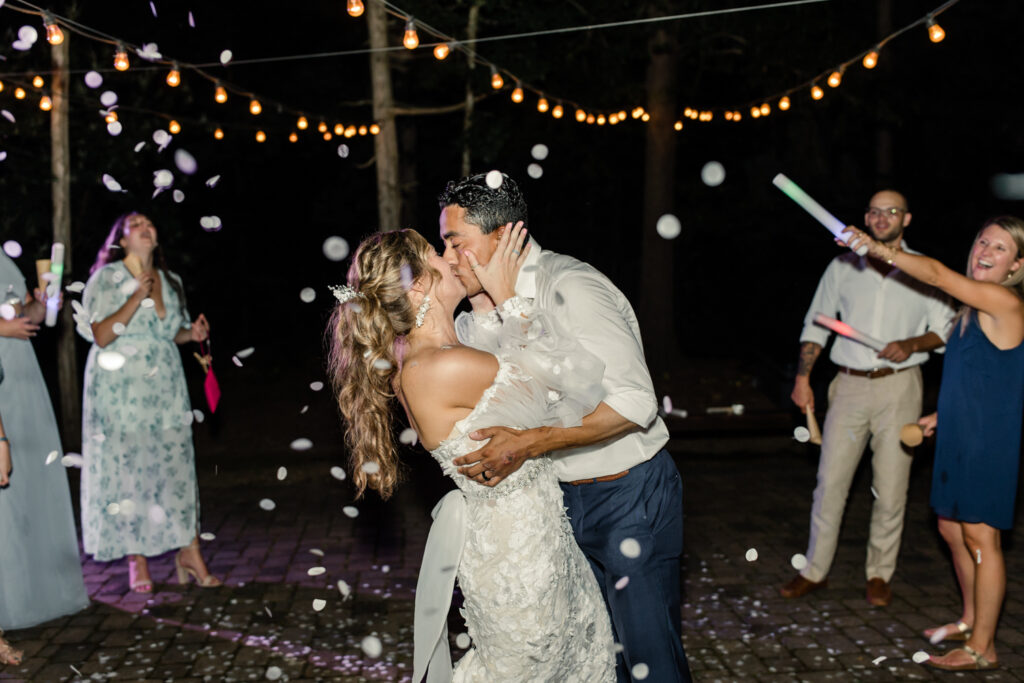Bride, in white lace dress, kisses groom, in white button down and blue pants, under confetti at night during reception exit at Morning Glory Farm Wedding Venue in Charlotte NC.