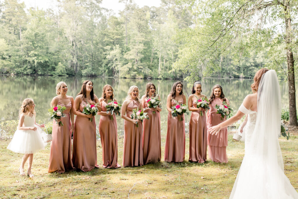 Eight bridesmaids, in pink dresses with pink bouquets, see bride, in white dress and veil, for the first time at The Millstone at Adams Pond Wedding Venue in Colombia SC