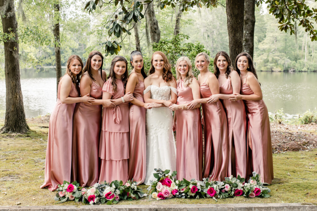 Eight bridesmaids, in pink dresses with pink bouquets, hugging bride, in white dress at The Millstone at Adams Pond Wedding Venue in Colombia SC