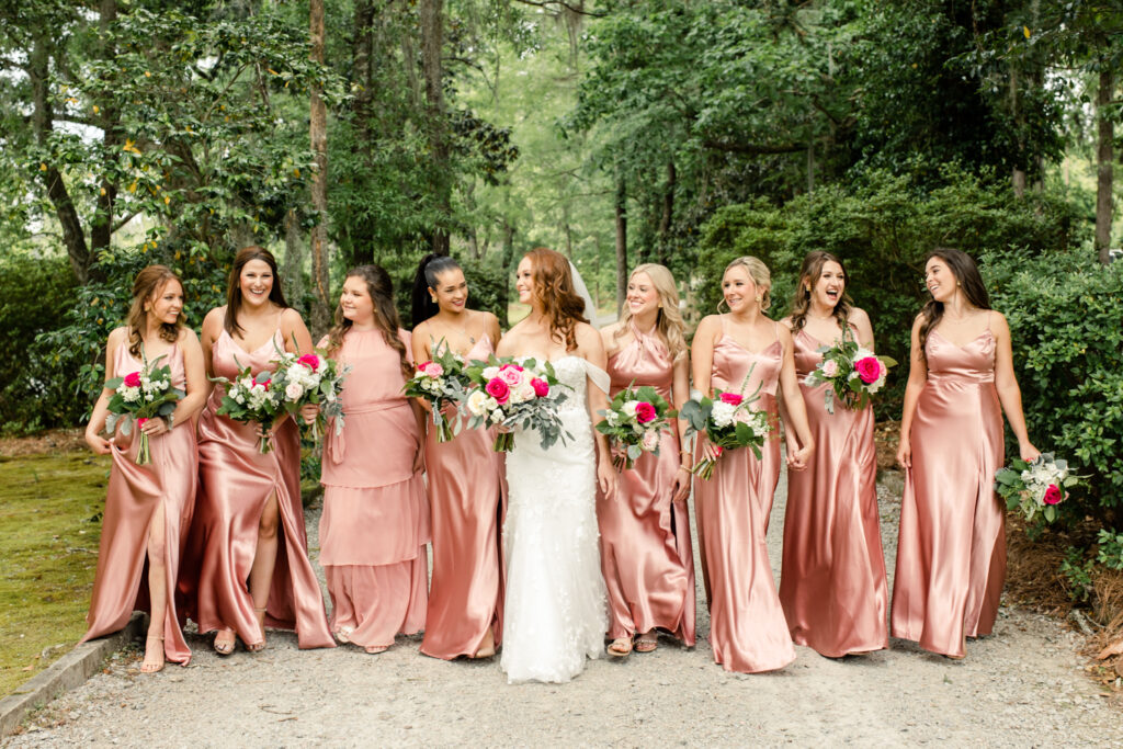 Eight bridesmaids, in pink dresses with pink bouquets, walking with bride, in white dress at The Millstone at Adams Pond Wedding Venue in Colombia SC
