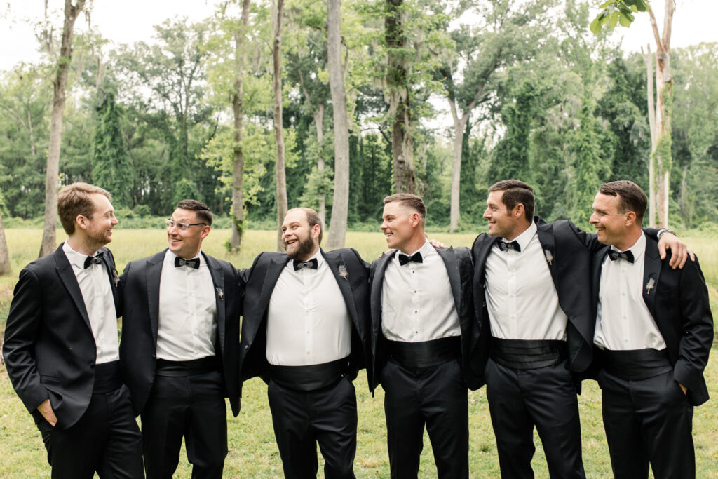 Five groomsmen, in white button down and black suites and bowties, laughing and hugging groom, in black suit at The Millstone at Adams Pond Wedding Venue in Colombia SC