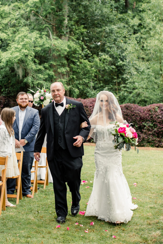 Bride, in white dress and veil holding pink bouquet, walks down aisle with father, in black suit, at The Millstone at Adams Pond Wedding Venue in Colombia SC