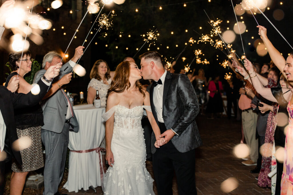 Bride, in white lace dress, holding hands and kissing groom, in black suit, during sparkler exit at The Millstone at Adams Pond Wedding Venue in Colombia SC