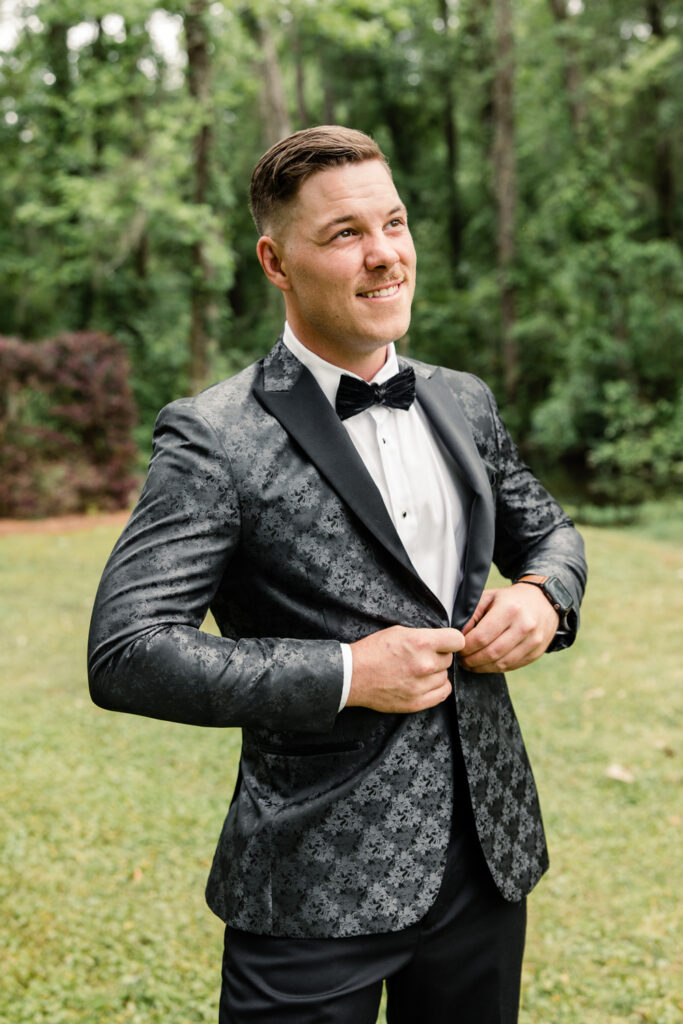 Groom buttoning up his black pattered suit with a black feather bowtie and white button down at The Millstone at Adams Pond Wedding Venue in Colombia SC