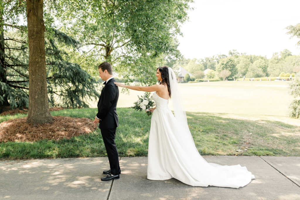 Bride, in white strapless gown and veil tapping left shoulder of groom, in black suit, during first look at Long View Country Club Wedding Venue Charlotte NC